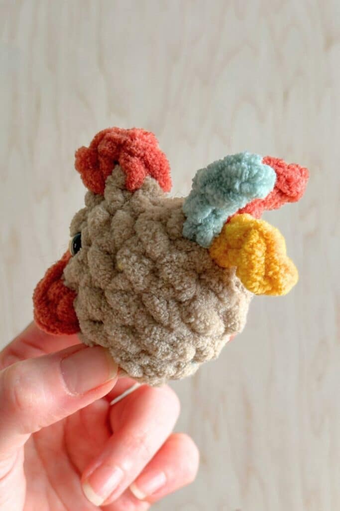 Mini crocheted rooster in a hand looking at back with different colored tail feathers.