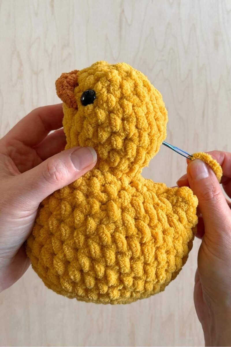 Hands holding a crochet suck with a yarn needle inserted at the back of the head. 