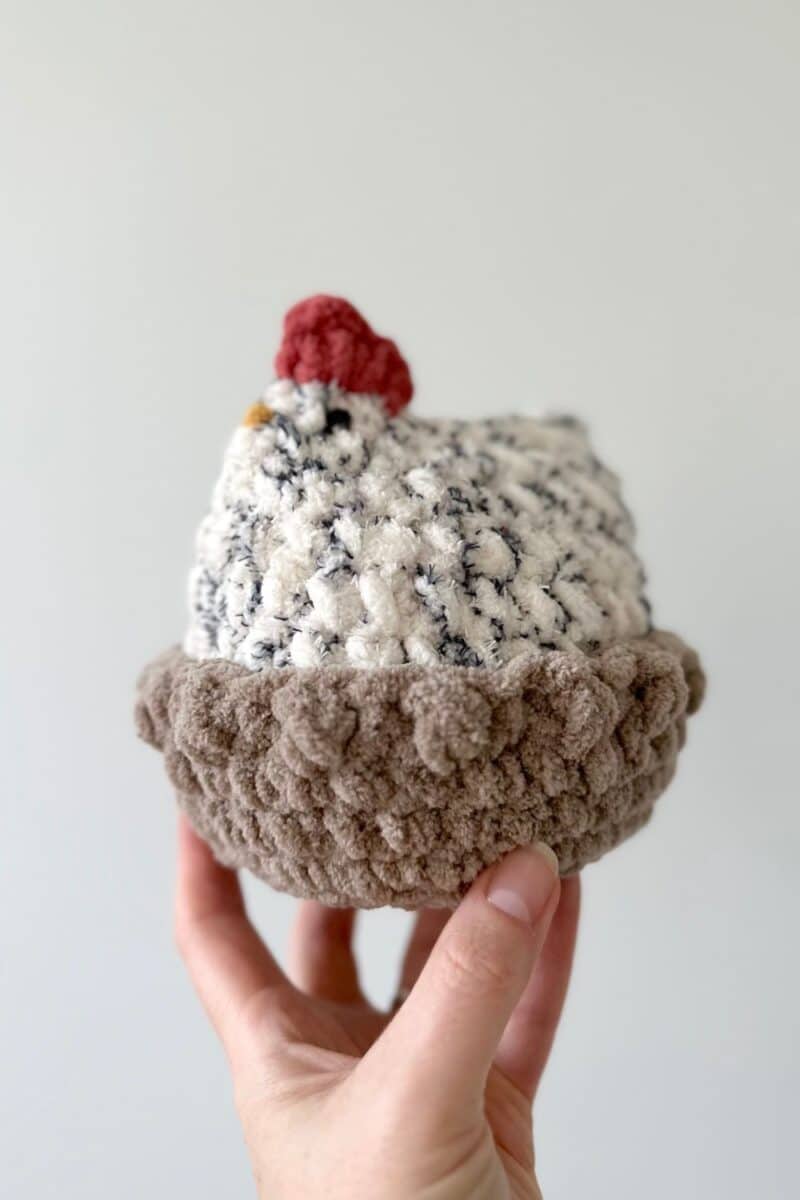 A hand holding a crochet chicken in a crochet nest.
