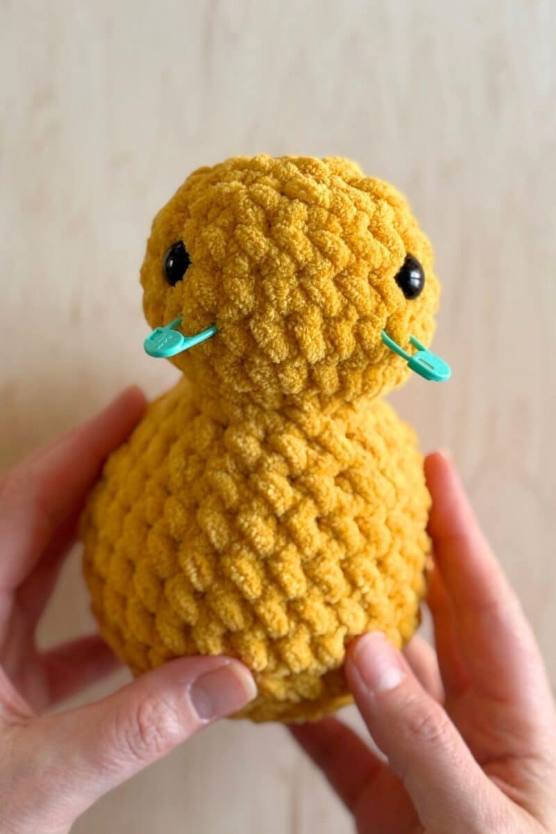 A process photo of a yellow crochet duck. Stitch markers are placed where the beak will go.