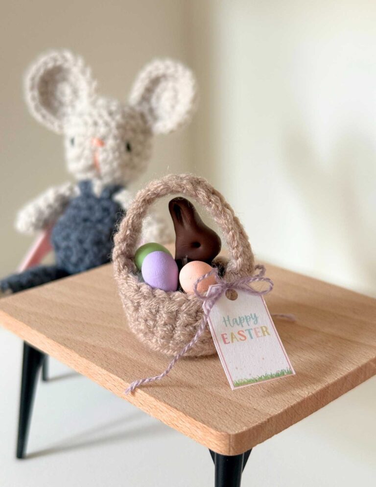 A tiny crochet dollhouse mouse sitting at a table with a dollhouse sized Easter basket with eggs, chocolate bunny and gift tag.