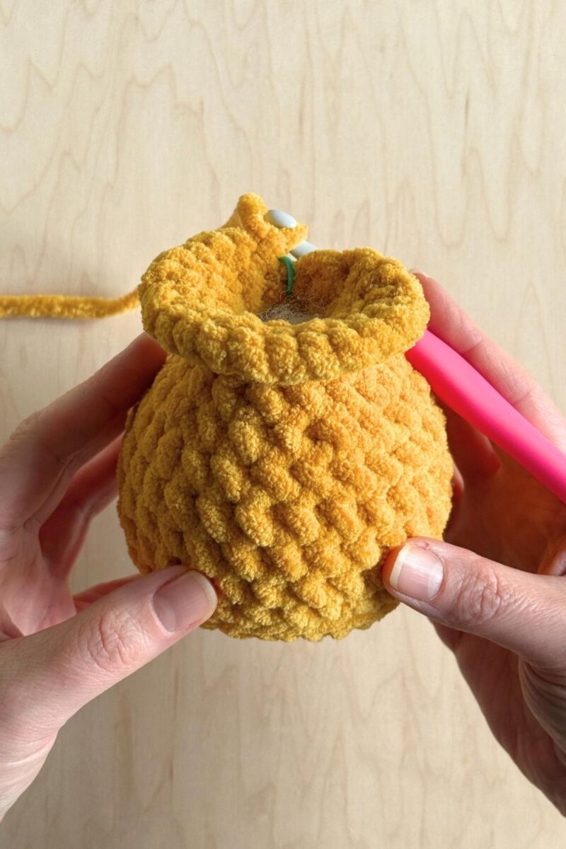 Hands holding a yellow crochet duck with the head partway completed.