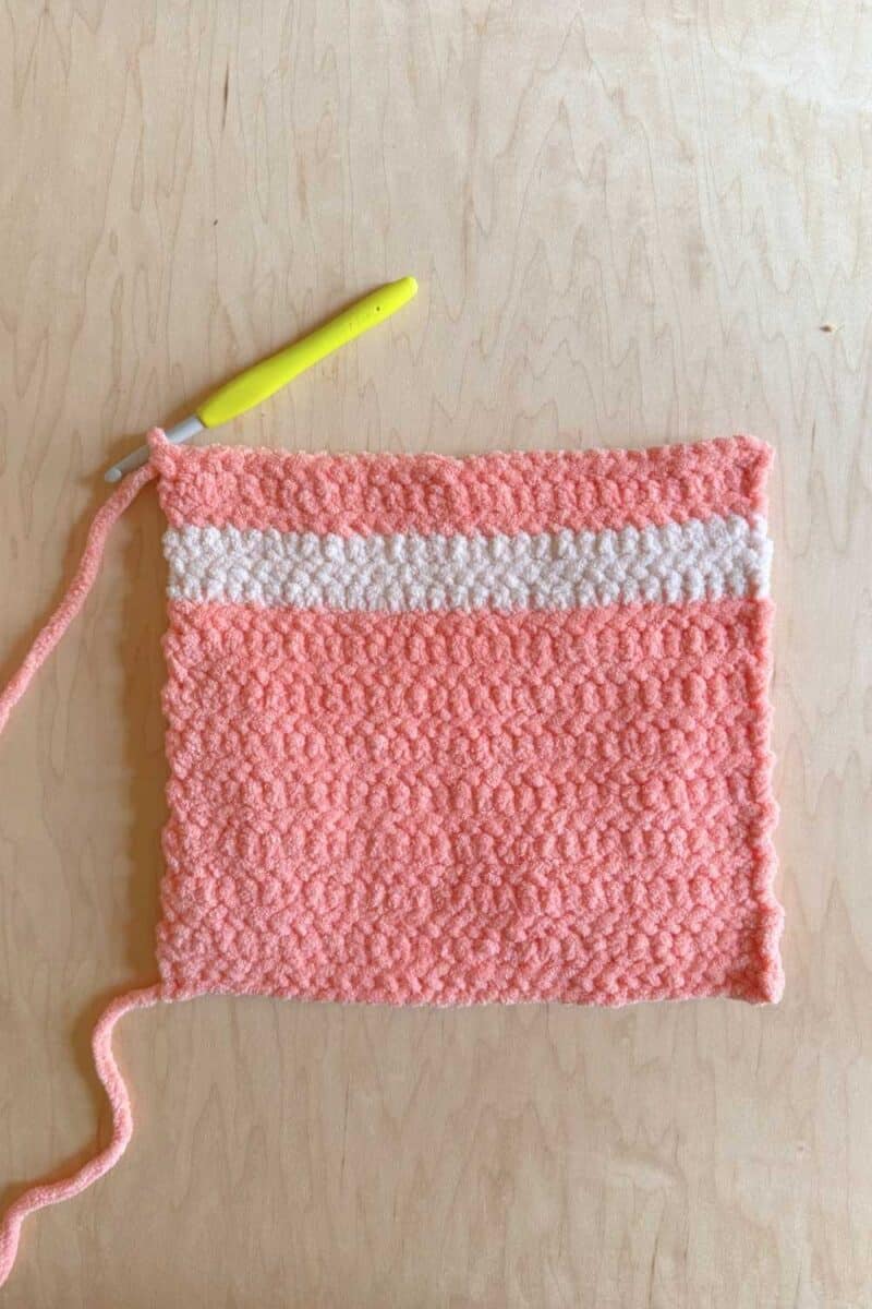 A pink crochet block with a light pink stripe. There is also a yellow crochet hook.