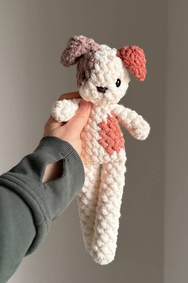 Hand holding a patchy crochet dog in neutral colors.