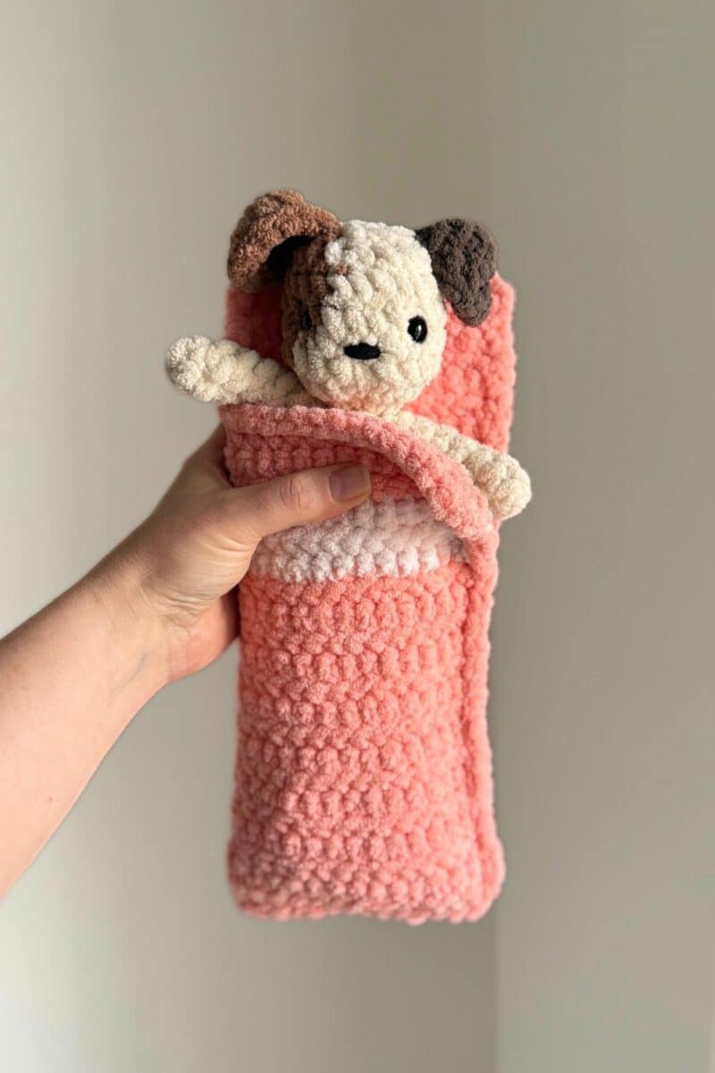 Hand holding a brown spotted crochet dog in a pink crochet sleeping bag.