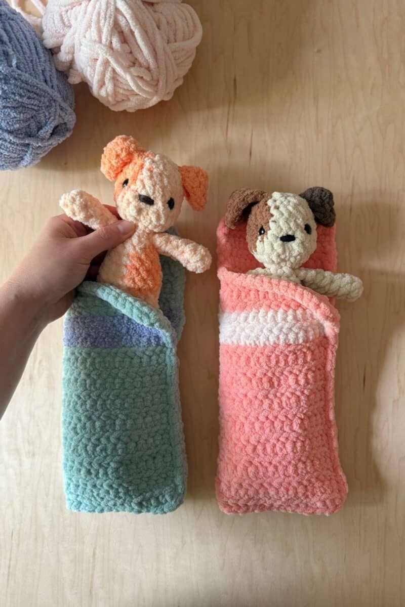 Two crochet sleeping bags in blue and pink with crochet dogs inside. There is a hand pulling one out.