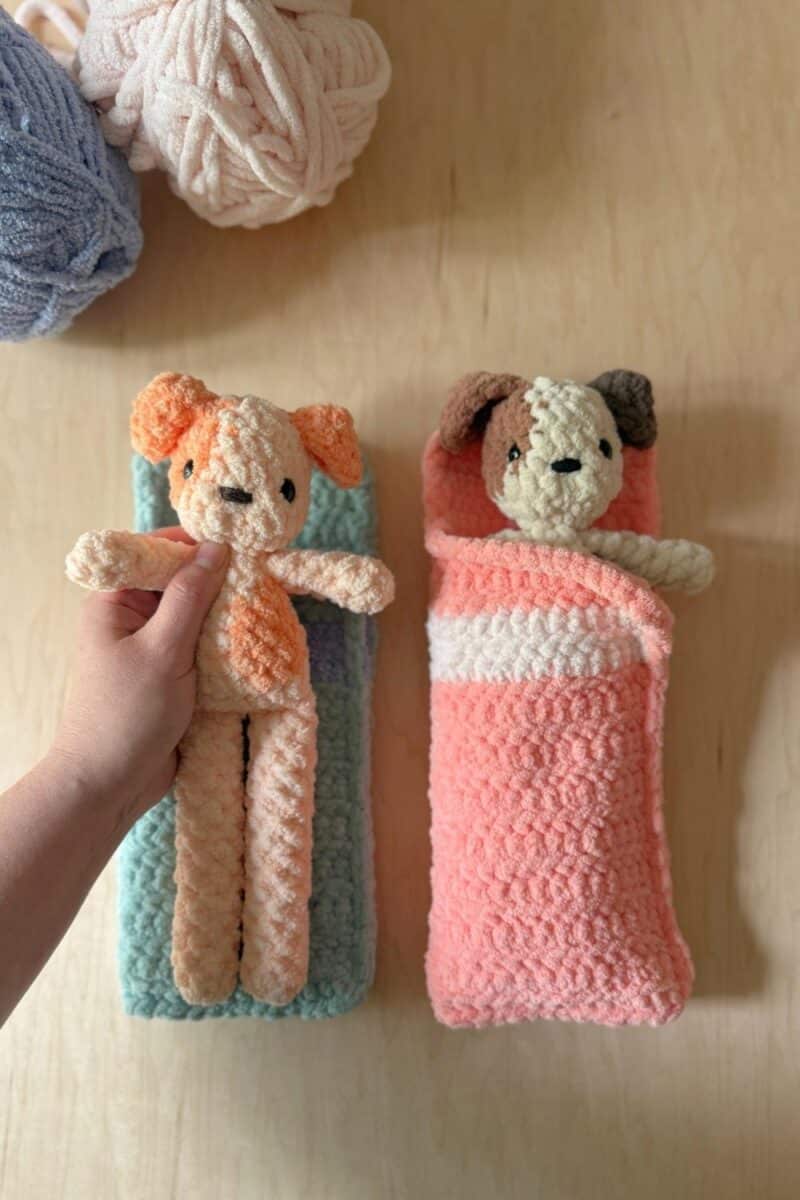Two crochet sleeping bags in blue and pink. One crochet dog is inside the pink sleeping bag and  a hand is holding one on top of the blue one. 