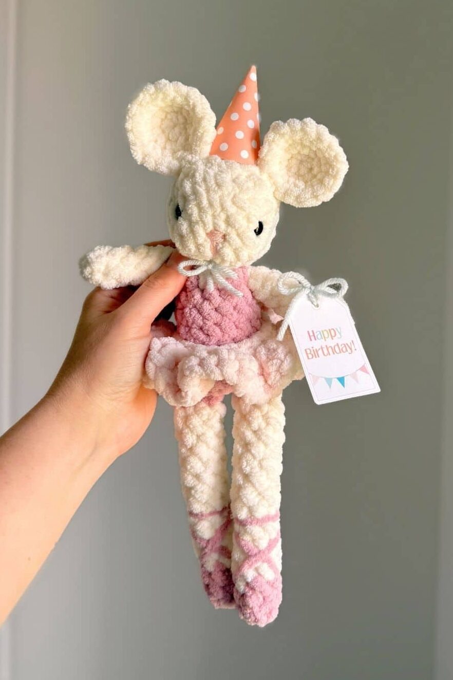 Hand holding a crochet ballet mouse wearing a paper birthday hat and tag.