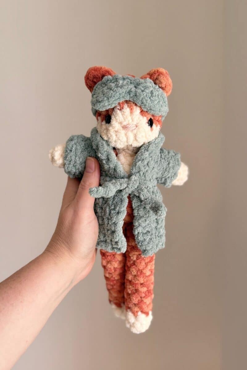 Hand holding an orange crochet cat wearing a robe and eye mask.