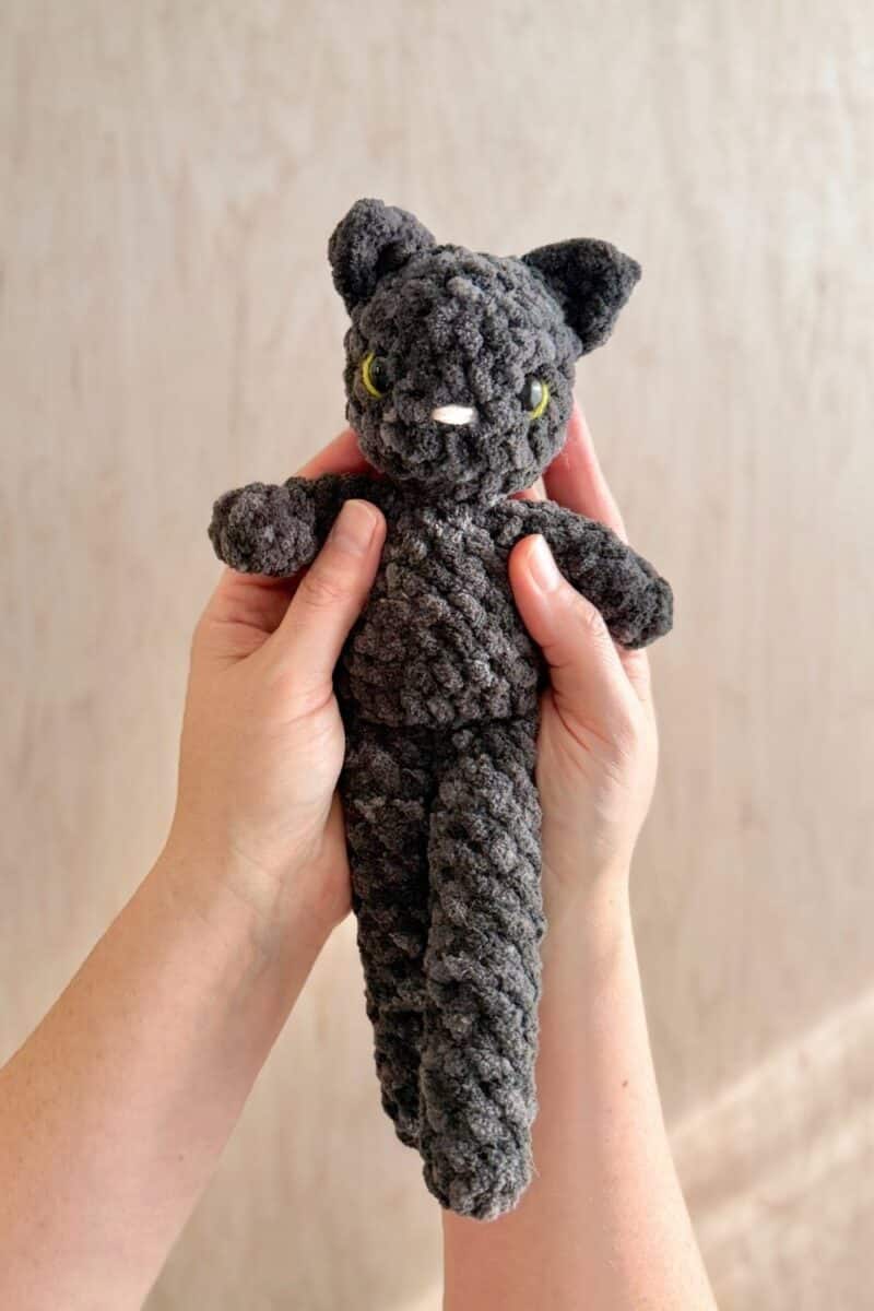 hands holding a dark grey/black crochet cat with green accents in her eyes.