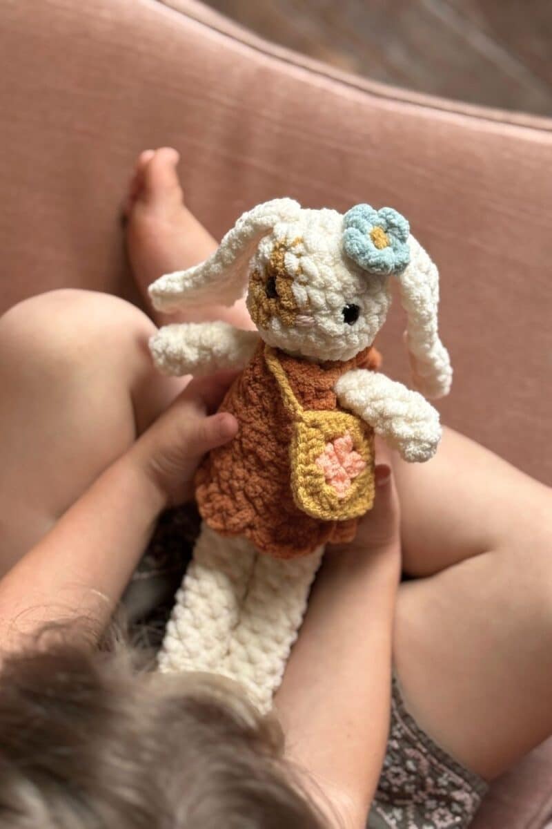 A little girl sitting on a chair form the top view. She is holding a little white bunny wearing a dress and a matching purse.