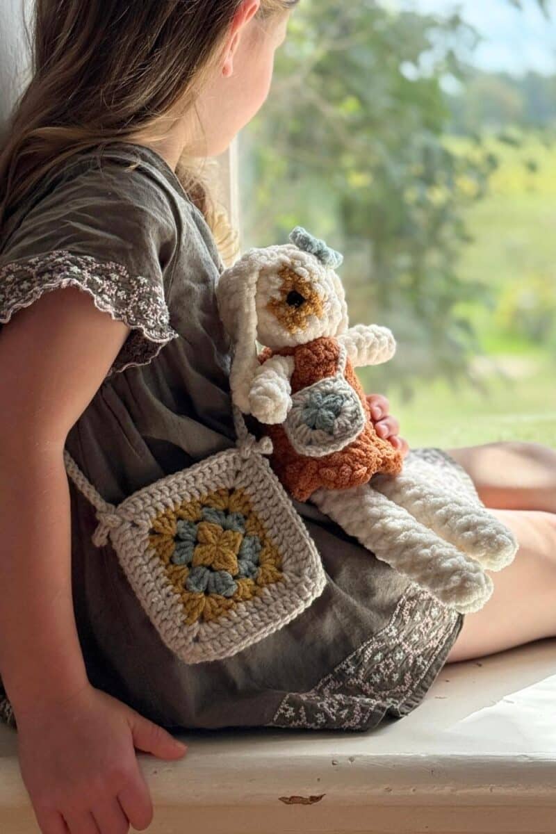 A little girl sitting in a window wearing a green dress and a crochet granny-square purse. She is holding a little white bunny wearing a dress and a matching purse.