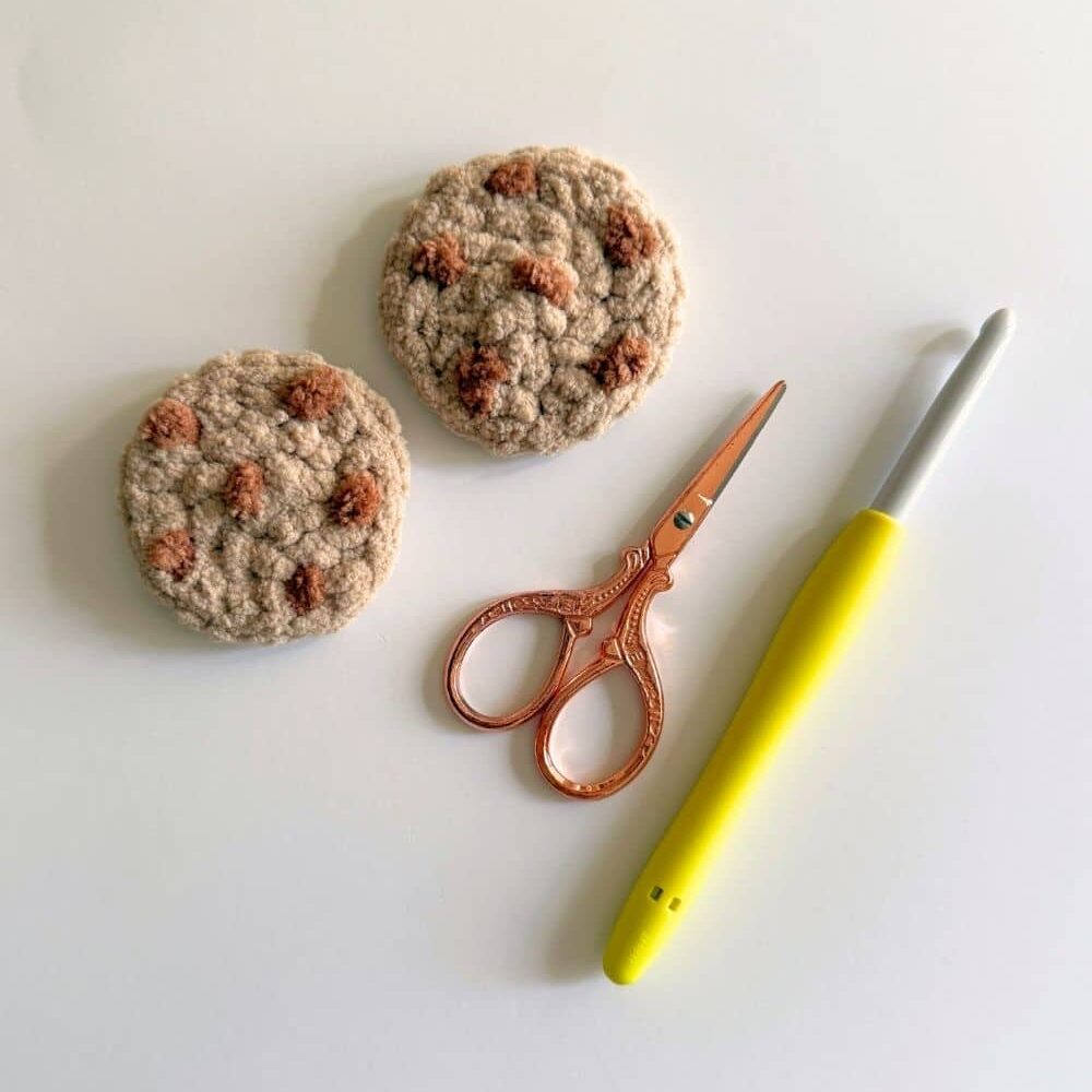 Two crochet cookies with scissors and a yellow crochet hook.
