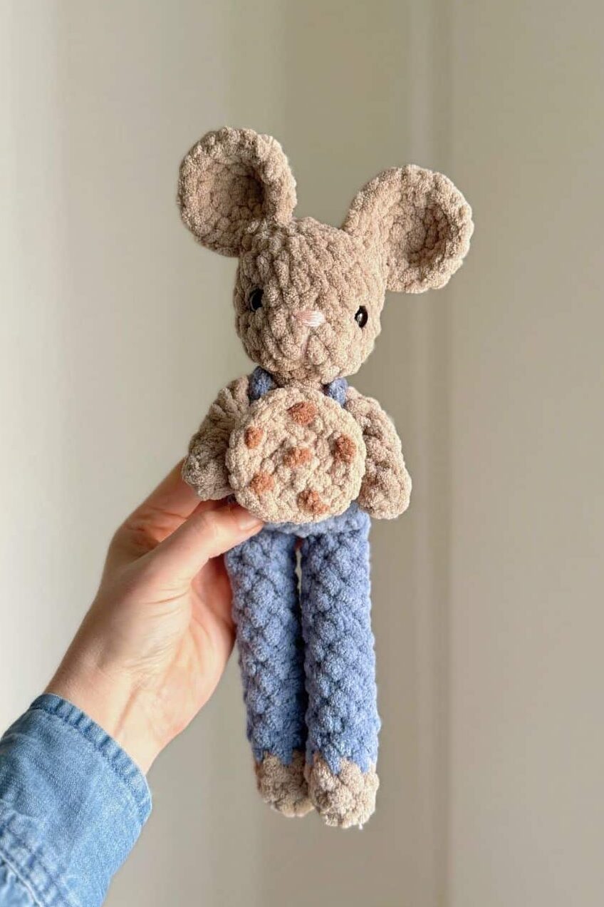 Hand holding a crochet mouse wearing blue overalls and holding a cookie.