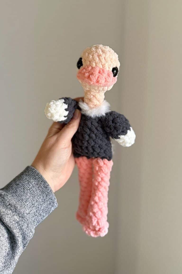 Hand holding a grey and pink crochet ostrich with white fur around her neck.