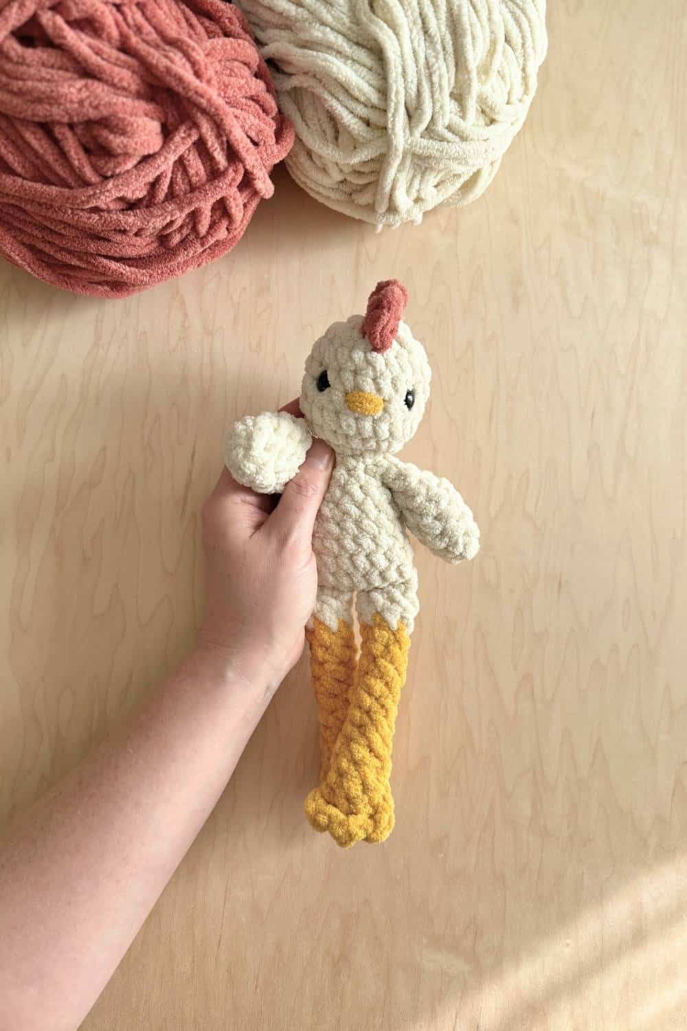 Hand holding a chicken crochet pattern on a wood surface with yarn in the top left corner.