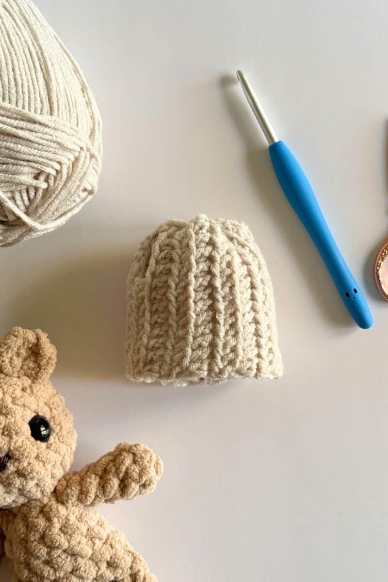 Photo showing a finished crochet beanie hat with crochet hook, yarn and a little bear face peeking into the frame.