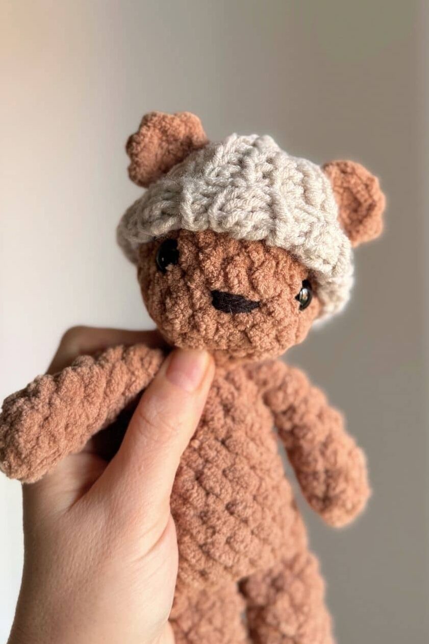 Hand holding a brown crochet bear wearing the finished hat.