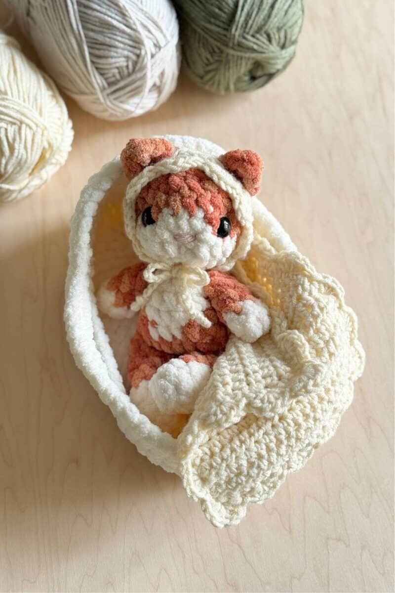 A baby crochet orange kitty in a bassinet with blanket and a bonnet on.