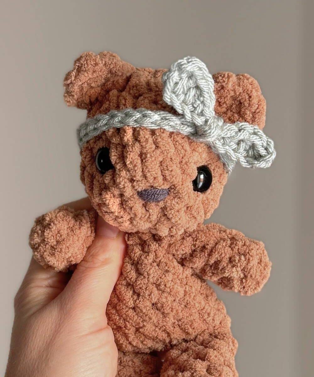 Hand holding a little crochet brown bear wearing a blue knotted headband.