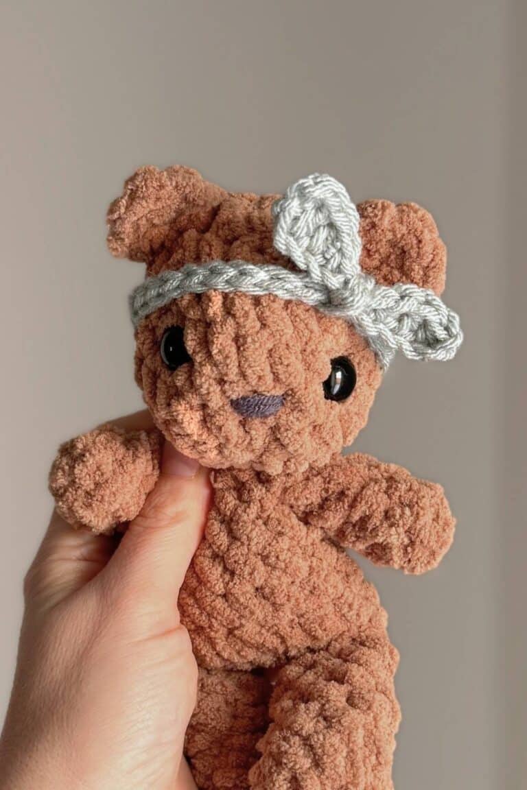 Hand holding a little crochet brown bear wearing a blue knotted headband.