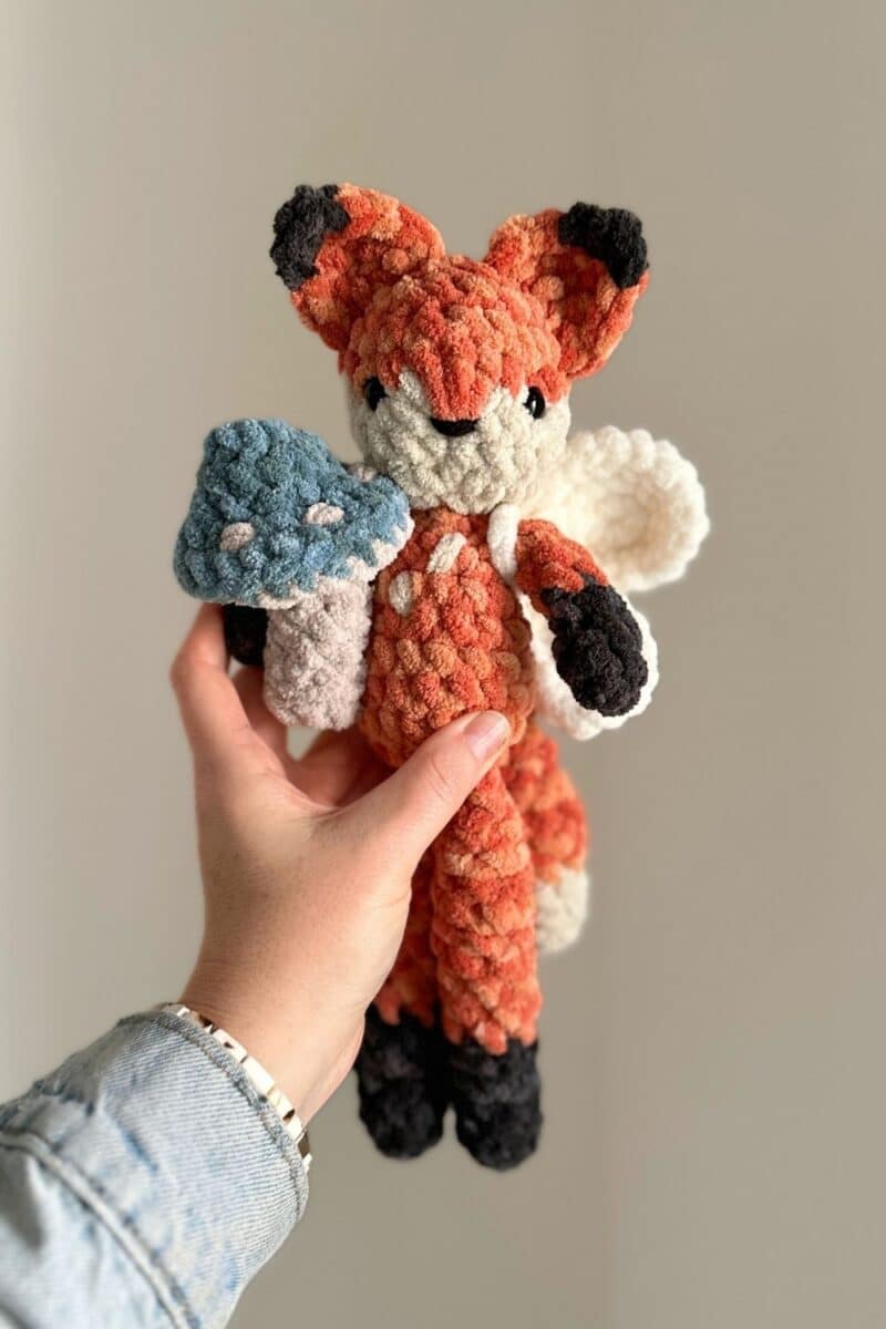 Hand holding a crochet fox - he is holding a little blue mushroom and wearing fairy wings.