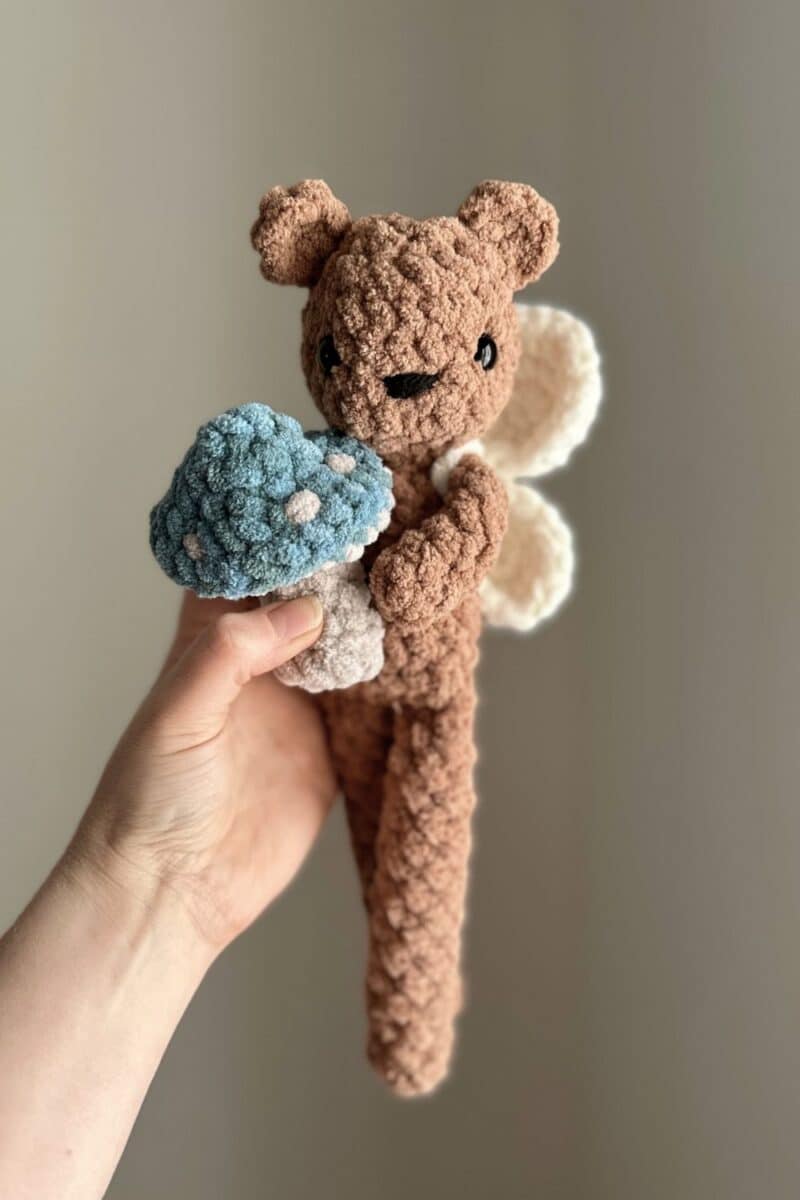 hand holding a brown crochet bear holding a mushroom and wearing fairy wings.