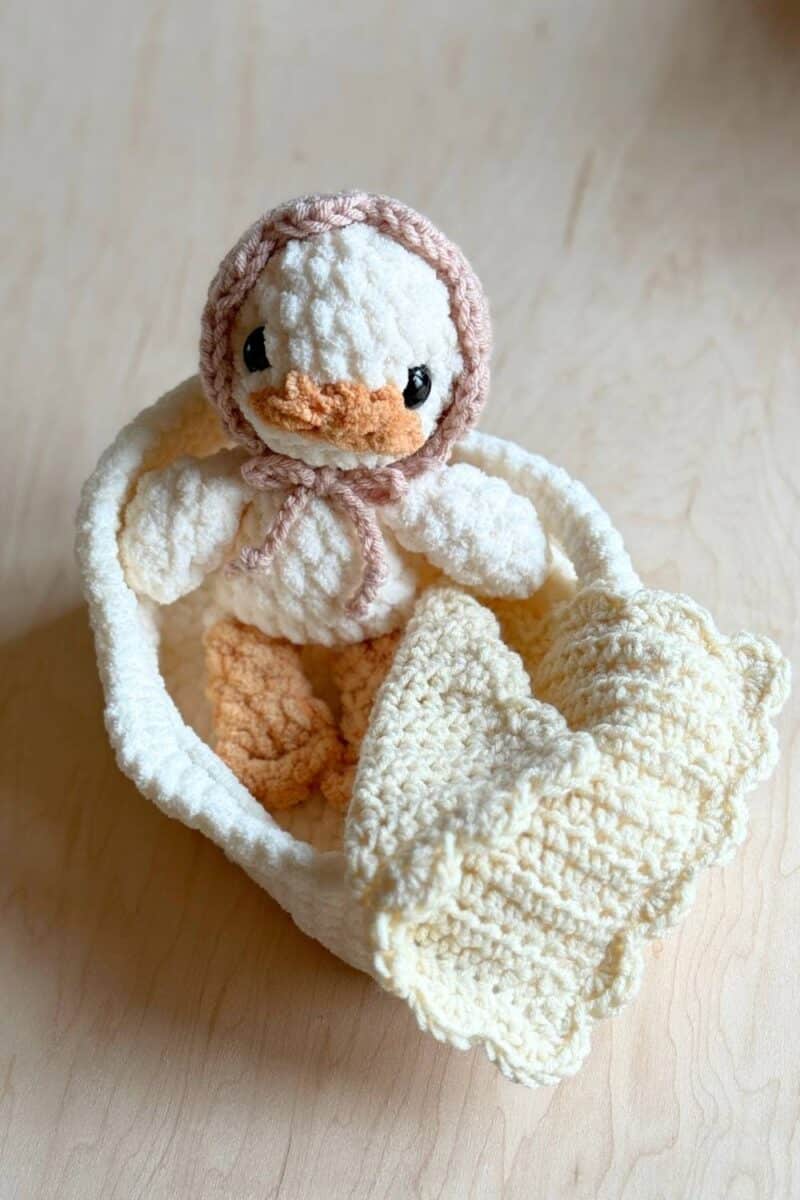 A crochet baby duck in a bassinet with blanket and bonnet.
