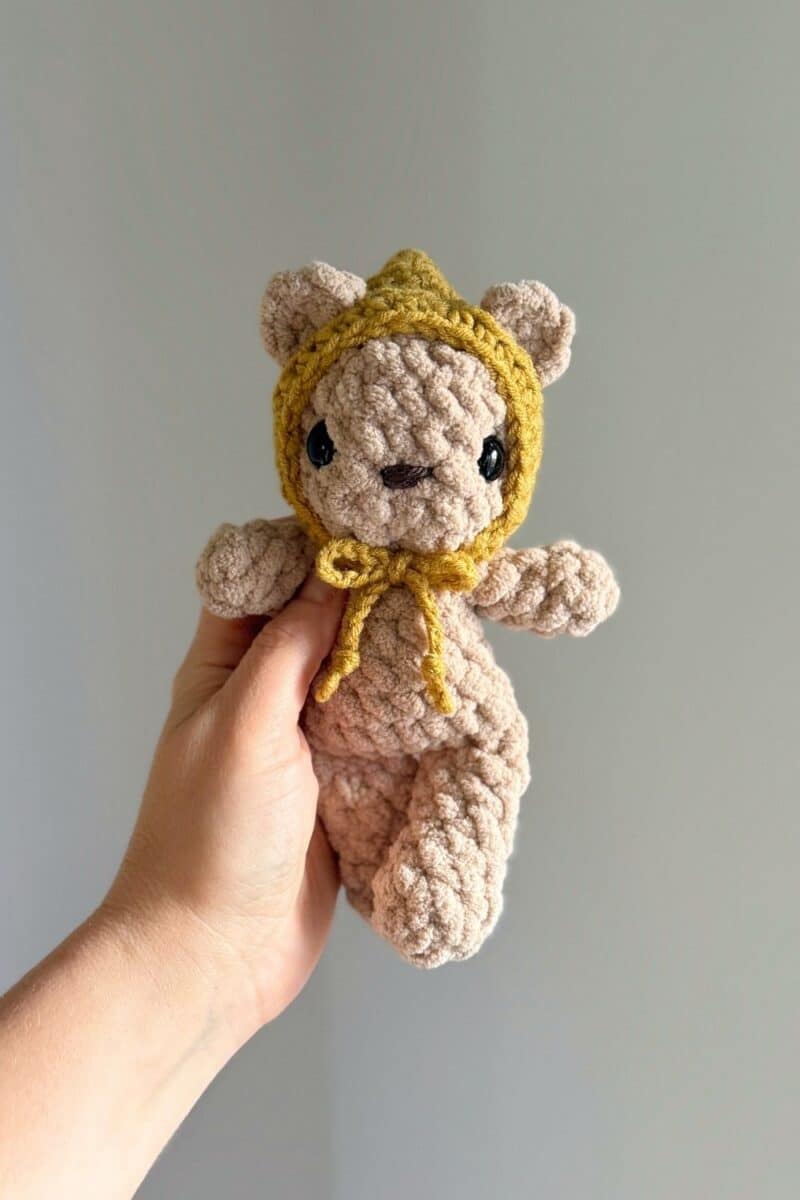 Hand holding a crochet brown baby bear wearing a yellow hat.