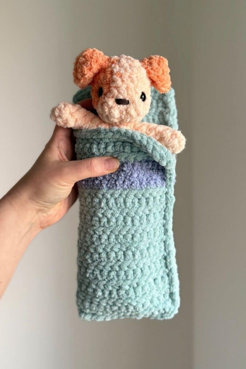Hand holding a crochet sleeping bag with an orange spotted crochet dog inside.