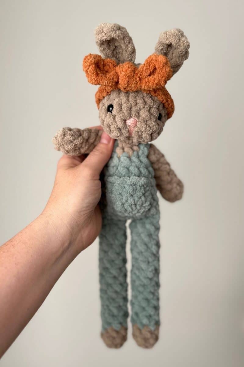 Hand holding a brown bunny wearing green overalls with an orange headband.
