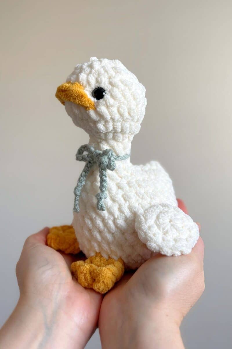 Hands holding a little white crochet goose with a blue crochet bow tied around her neck.