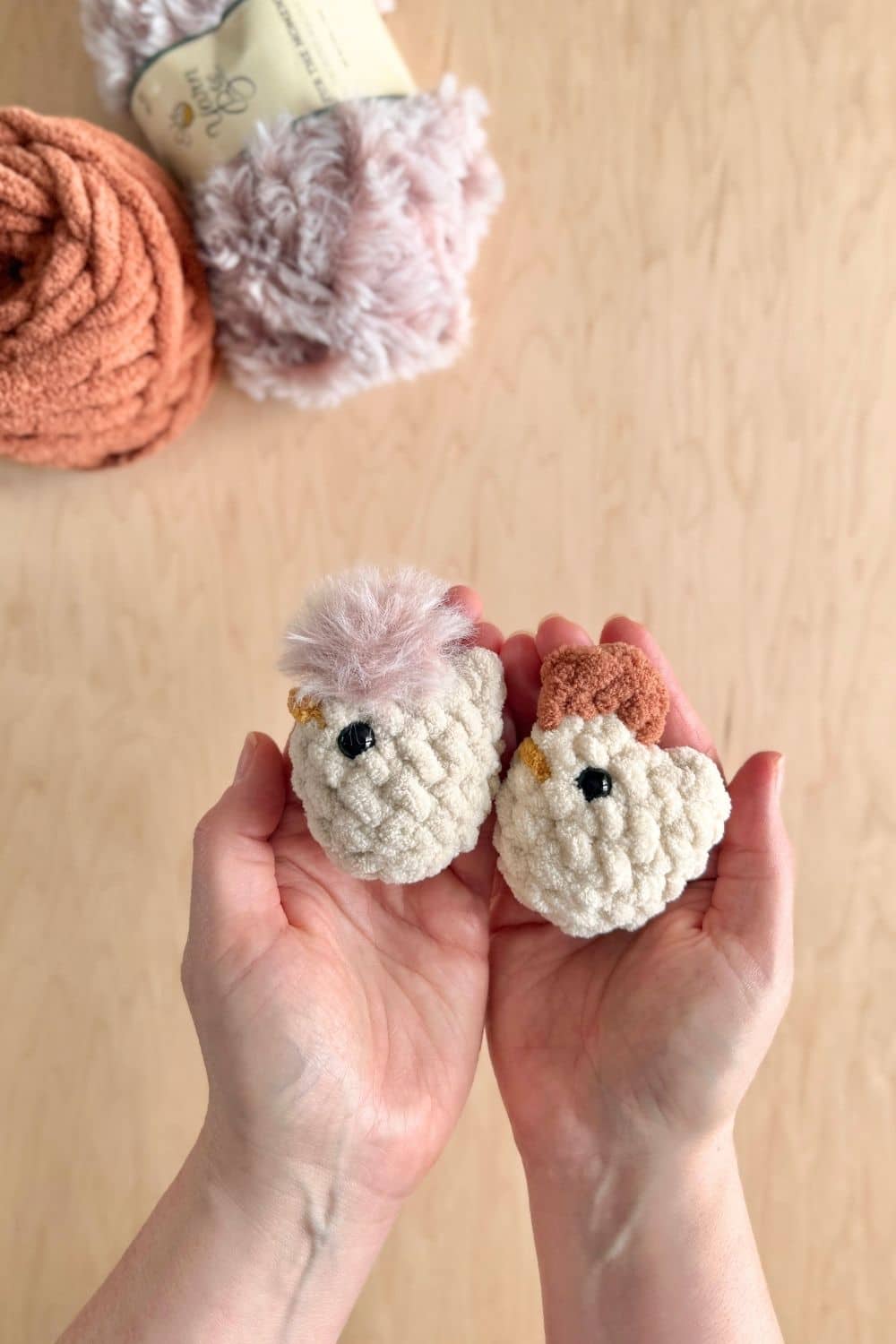Hands holding two mini crochet chickens with yarn in the background.