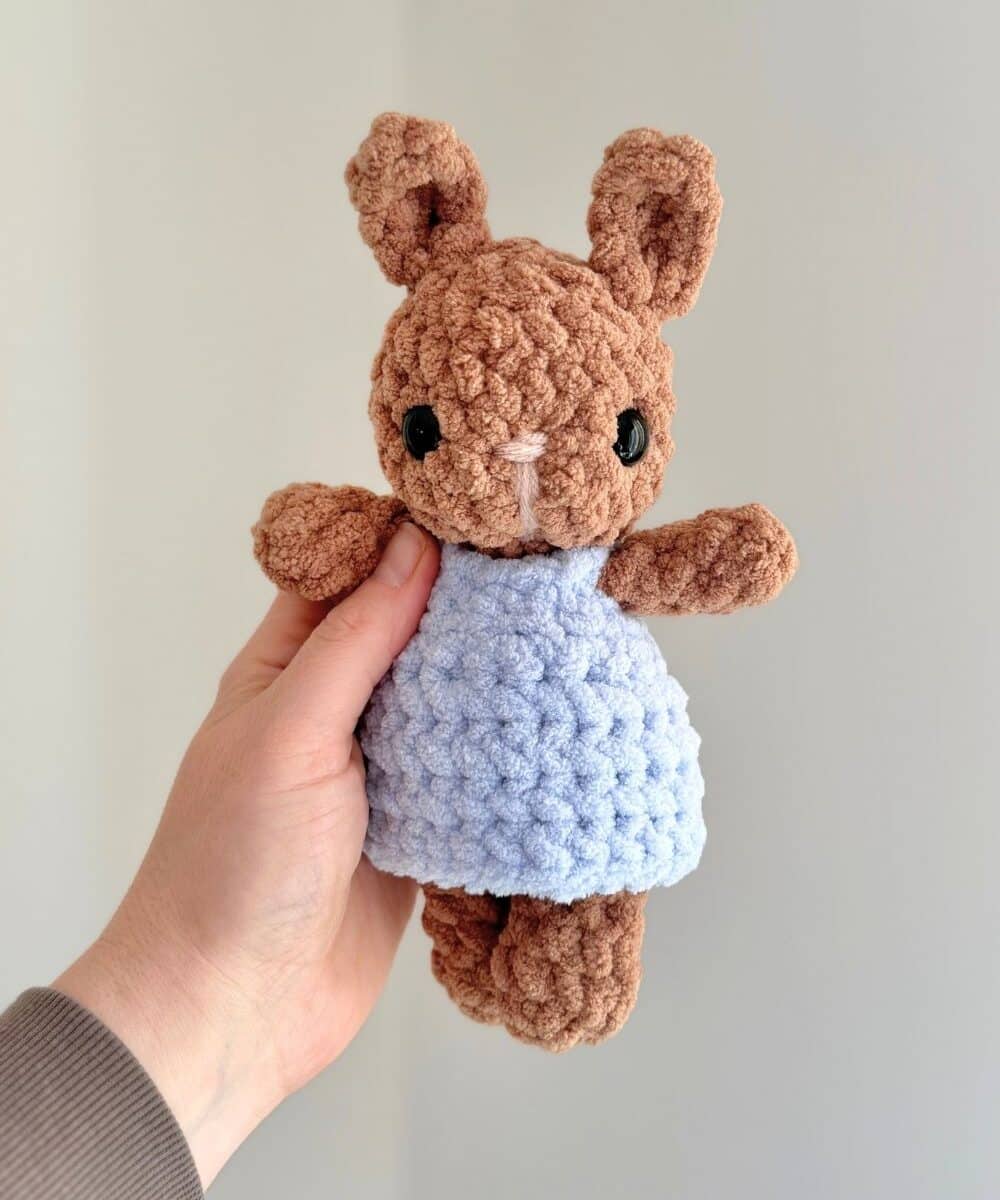Hand holding a brown baby crochet bunny wearing a blue crochet dress.