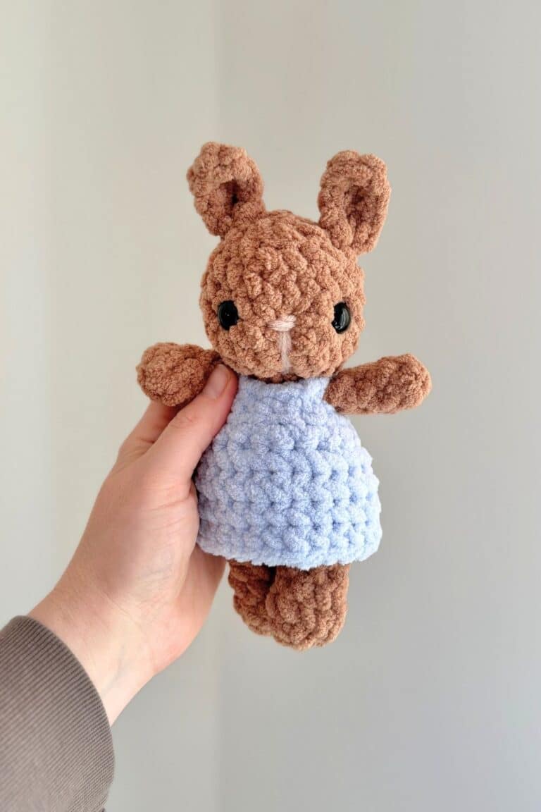 Hand holding a brown baby crochet bunny wearing a blue crochet dress.