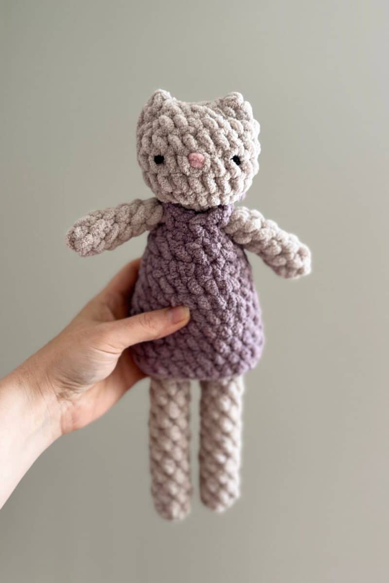 A grey crochet cat wearing a purple dress.