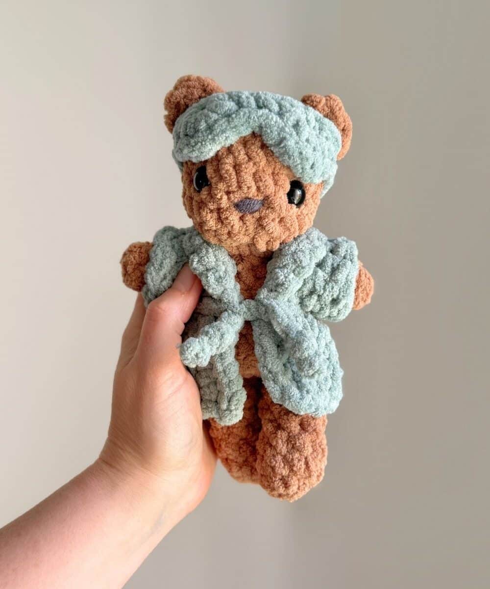 A crochet teddy bear wearing a crochet robe and eye mask.
