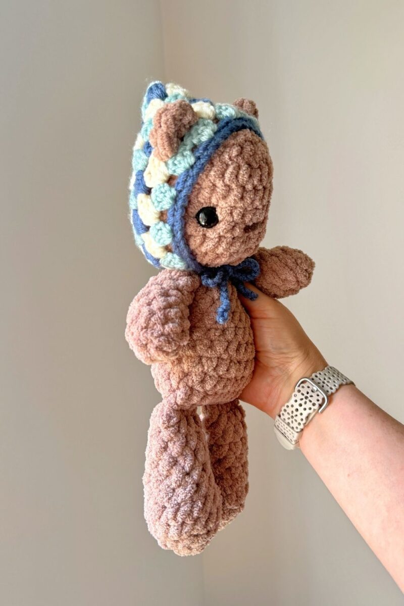 Hand holding a light brown crochet bear that's wearing a crochet vintage style pixie hat that's been crocheted as well.
