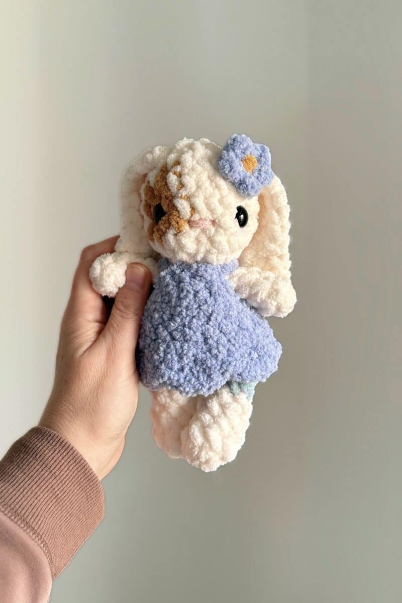 A tiny crochet bunny with long ears wearing a dress and flower.